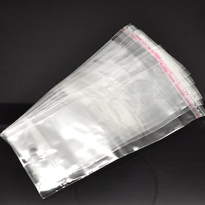 May include: A stack of clear cellophane bags with a red strip at the top edge. The red strip has white text that reads "REMOVE LINER TO EXPOSE SEALING TAPE". The bags are transparent and appear to be empty.