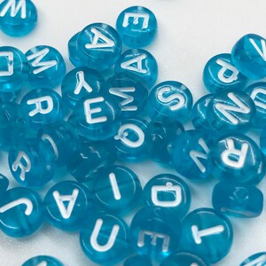 Może przedstawiać: Zbi&oacute;r niebieskich plastikowych koralik&oacute;w alfabetycznych z białymi literami. Koraliki są rozrzucone na białym tle.