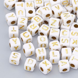 May include: White plastic letter beads with gold lettering. The beads are in the shape of cubes and feature all the letters of the alphabet.
