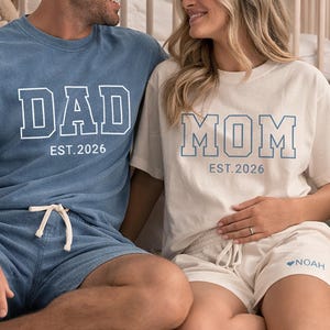 Peut inclure: Un couple porte des ensembles assortis de t-shirts et shorts bleus et cr&egrave;me. Le t-shirt bleu dit "DAD EST. 2026" et le t-shirt cr&egrave;me dit "MOM EST. 2026". Des chaussures de b&eacute;b&eacute; et des blocs &eacute;pellent "BABY". Un ours en peluche est &agrave; proximit&eacute;.