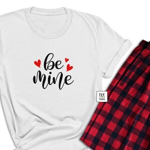 May include: A white t-shirt with the words "be mine" in black script and red heart accents, paired with red and black plaid pajama pants. The pajama pants have a drawstring closure.