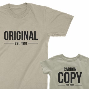 May include: Two beige t-shirts with black text. The larger shirt says "ORIGINAL - EST. 1991". The smaller shirt says "CARBON COPY - EST. 2023".