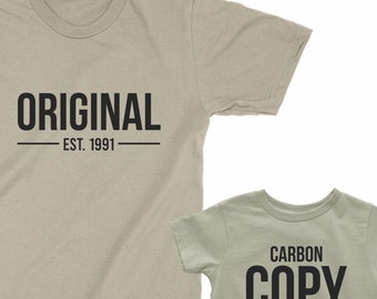 May include: Two beige t-shirts with black text. The larger shirt says "ORIGINAL - EST. 1991". The smaller shirt says "CARBON COPY - EST. 2023".