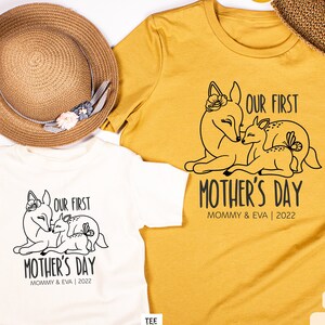 Peut inclure: Deux t-shirts, un blanc et un jaune moutarde, avec un motif noir représentant une biche mère et son faon, accompagnés du texte "Our First Mother's Day Mommy & Eva | 2022"