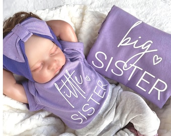 May include: A purple baby onesie with the text "little sister" and a heart. A purple shirt with the text "big sister" and a heart. The baby is wearing a purple headband.