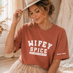 May include: A rust-colored short-sleeve t-shirt featuring the white text WIFEY SPICE AND EVERYTHING NICE on the front and a small heart icon with the text EST. 2026 on the sleeve, paired with a beige fedora hat and a long cream-colored lace skirt.