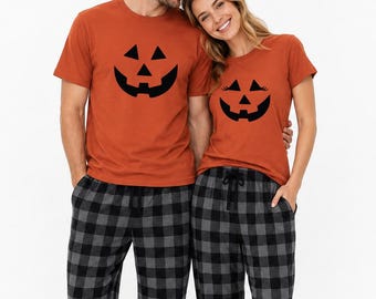 May include: Two matching sets of pajamas consisting of burnt orange short-sleeved t-shirts with black jack-o'-lantern face graphics and grey and black buffalo plaid flannel pajama pants with an elastic drawstring waistband.