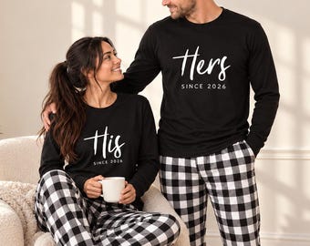May include: Matching black long-sleeve shirts with white text reading His since 2026 and Hers since 2026, paired with black and white buffalo plaid pajama pants. The shirts feature a crew neckline and the pants have a classic checkered pattern.