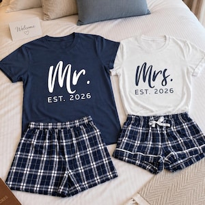 Pu&ograve; includere: Una t-shirt blu navy con testo bianco Mr. EST. 2026 e una t-shirt bianca con testo blu navy Mrs. EST. 2026, abbinate a due paia di pantaloncini a quadri blu navy e bianchi con elastico in vita e coulisse bianca.