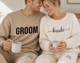 May include: Matching beige loungewear sets with the words "GROOM" and "bride" printed on the tops. The bottoms have "EST. 2026" printed on them. The couple is holding mugs. The bride's top is cream-colored, and the groom's is tan.