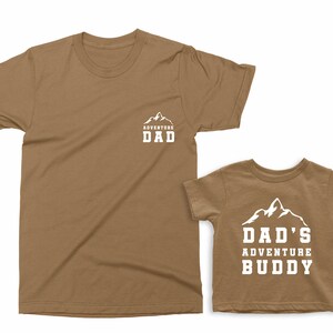 May include: Two brown t-shirts, one adult and one child size. The adult shirt has "ADVENTURE DAD" printed in white with a mountain graphic. The child's shirt says "DAD'S ADVENTURE BUDDY" with a mountain graphic.