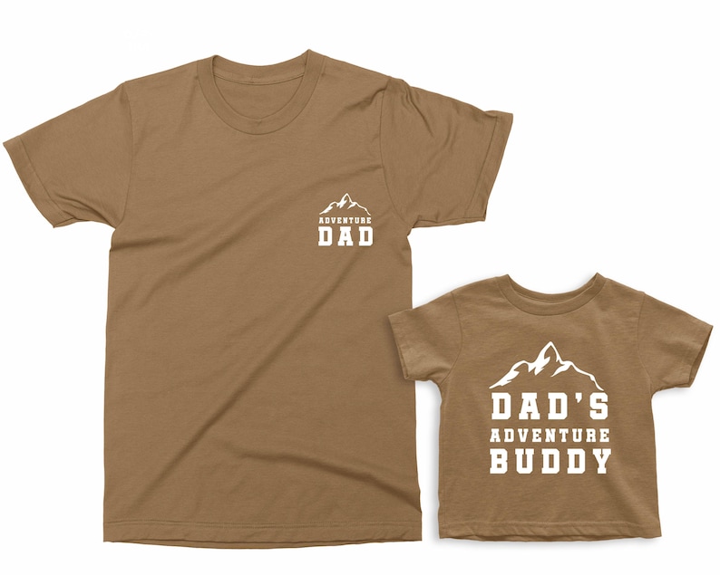 May include: Two brown t-shirts, one adult and one child size. The adult shirt has "ADVENTURE DAD" printed in white with a mountain graphic. The child's shirt says "DAD'S ADVENTURE BUDDY" with a mountain graphic.