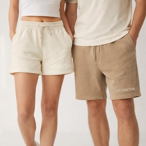 May include: Cream-colored and tan shorts with the text "THE SMITHS" printed in white. The shorts have a drawstring waist and pockets. The shorts are made of a soft material.