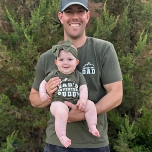 May include: A man wearing a green t-shirt with the text "ADVENTURE DAD" holds a baby wearing a matching green outfit. The baby's outfit says "DAD'S ADVENTURE BUDDY". Both are smiling in front of green foliage.