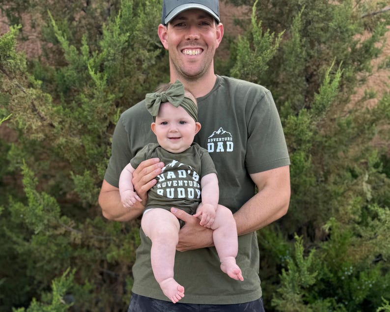 May include: A man wearing a green t-shirt with the text "ADVENTURE DAD" holds a baby wearing a matching green outfit. The baby's outfit says "DAD'S ADVENTURE BUDDY". Both are smiling in front of green foliage.