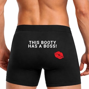 May include: Black boxer briefs with the text "THIS BOOTY HAS A BOSS!" in white. A red lip print is on the right side. The briefs are made of a soft material.