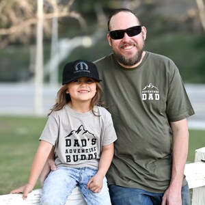 May include: A father and child pose for a photo. The child wears a gray t-shirt with the text "DAD'S ADVENTURE BUDDY" and a black cap. The father wears a green t-shirt with the text "ADVENTURE DAD".