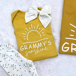 Może przedstawiać: Ż&oacute;łty body z białym nadrukiem słońca i tekstem "Grammy's Sunshine". Na body znajduje się biała kokardka.