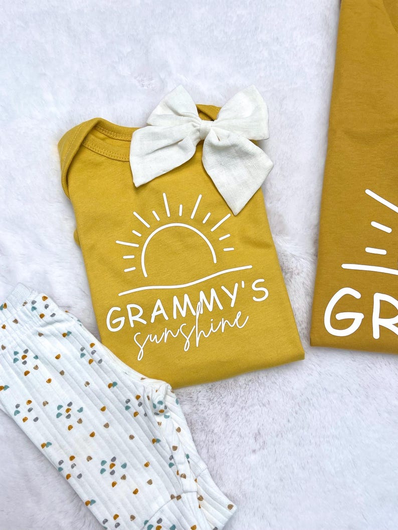 Może przedstawiać: Ż&oacute;łty body z białym nadrukiem słońca i tekstem "Grammy's Sunshine". Na body znajduje się biała kokardka.