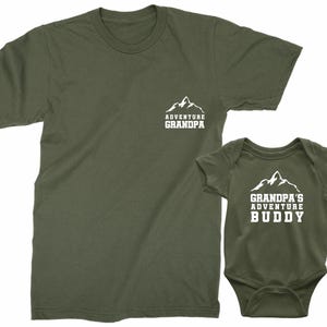 May include: A green t-shirt with the text "Adventure Grandpa" and a mountain graphic. A matching green baby onesie with the text "Grandpa's Adventure Buddy" and a mountain graphic.