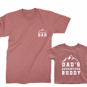 May include: Two matching dusty rose t-shirts. One adult tee has a mountain graphic with the text "ADVENTURE DAD". The other is a child's tee with the text "DAD'S ADVENTURE BUDDY" and a mountain graphic.