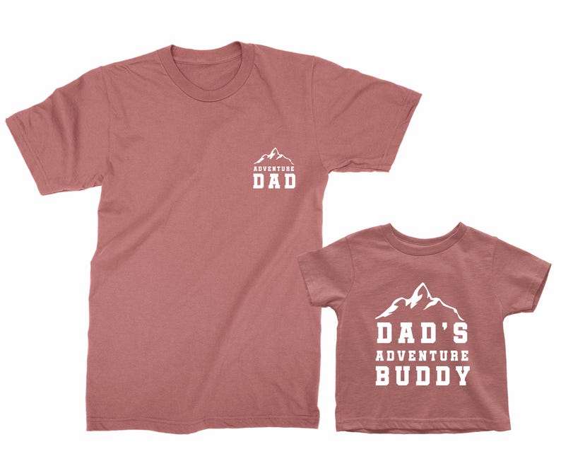 May include: Two matching dusty rose t-shirts. One adult tee has a mountain graphic with the text "ADVENTURE DAD". The other is a child's tee with the text "DAD'S ADVENTURE BUDDY" and a mountain graphic.