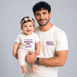 May include: A man and a baby wearing matching cream-colored t-shirts. The baby's shirt says "Tough Dad? Watch Me Smile" in purple text. The man's shirt says "Built Tough...Until She Smiles". The baby wears a beige bow headband.