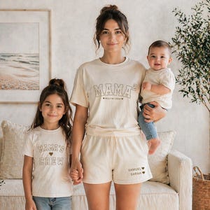 May include: A family of three wearing matching cream-colored t-shirts and shorts. The shirts read "MAMA" and "MAMA'S CREW" in a retro font. The shorts are printed with names. The family is standing in front of a neutral-toned background.