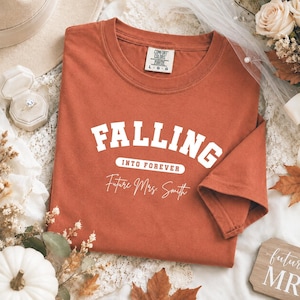 May include: A rust-colored t-shirt featuring white text that reads FALLING INTO FOREVER and Future Mrs. Smith. The shirt has a crew neckline and a Comfort Colors brand label on the inside collar. The fabric has a soft, washed appearance.