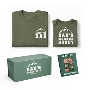 May include: Olive green t-shirt and baby onesie with white mountain graphic and text "Adventure Dad" and "Dad's Adventure Buddy". A matching gift box and photo frame complete the set. The photo frame has a picture of a man and a baby.