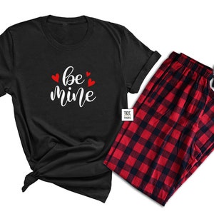 May include: A black t-shirt with the words "be mine" in white script and red heart accents, paired with red and black plaid pajama pants. The t-shirt is knotted at the bottom. The pajama pants have a drawstring.