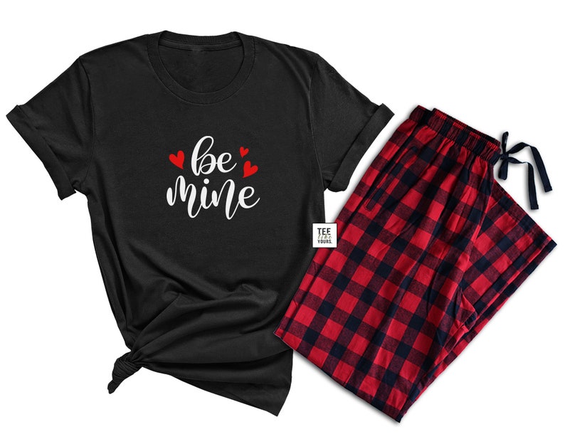 May include: A black t-shirt with the words "be mine" in white script and red heart accents, paired with red and black plaid pajama pants. The t-shirt is knotted at the bottom. The pajama pants have a drawstring.