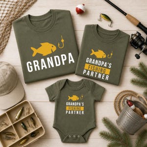 May include: Olive green t-shirts and a onesie with fishing-themed graphics. The shirts read "GRANDPA" and "GRANDPA'S FISHING PARTNER" with a yellow fish and hook design. Fishing gear and accessories surround the shirts.