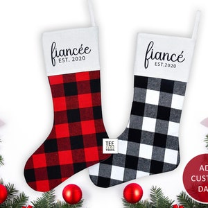 May include: Two red and white buffalo plaid Christmas stockings with white felt tops. The top of the stocking on the left says "fiancée EST. 2020" and the top of the stocking on the right says "fiancé EST. 2020".