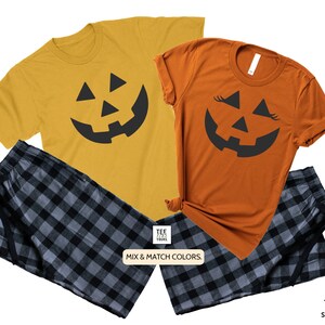 May include: A matching set of two t-shirts with a black jack-o'-lantern face design. One shirt is orange and the other is yellow. The shirts are paired with black and gray plaid pajama pants.