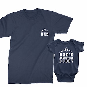 May include: Navy blue t-shirt with the text "ADVENTURE DAD" and a matching navy blue baby bodysuit with the text "DAD'S ADVENTURE BUDDY". Both items feature a white mountain graphic.