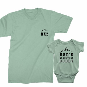 May include: Two matching light green shirts. One is a t-shirt with "ADVENTURE DAD" printed in black. The other is a baby onesie with "DAD'S ADVENTURE BUDDY" printed in black. Both shirts have a mountain graphic.