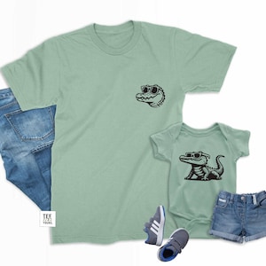 May include: Matching green t-shirts with a black alligator wearing sunglasses graphic. The adult shirt has the alligator's head only, while the baby shirt has the full alligator body.