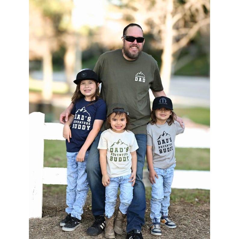 May include: A family of five poses outdoors, wearing matching apparel. The father wears a green t-shirt with "DAD" and a mountain graphic. The children wear t-shirts and hats with "DAD'S ADVENTURE BUDDY" text. All are wearing jeans.