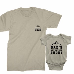May include: Tan t-shirt with the text "ADVENTURE DAD" and a matching baby onesie with the text "DAD'S ADVENTURE BUDDY". Both garments feature a mountain graphic in black.