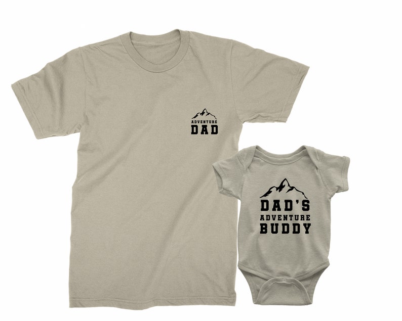 May include: Tan t-shirt with the text "ADVENTURE DAD" and a matching baby onesie with the text "DAD'S ADVENTURE BUDDY". Both garments feature a mountain graphic in black.