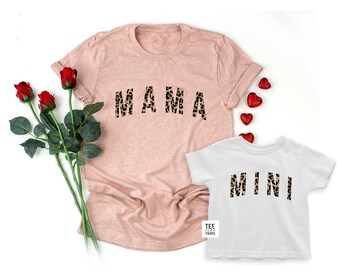 May include: Two pink and white t-shirts with a leopard print design. The larger shirt says "MAMA" and the smaller shirt says "MINI".