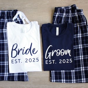 May include: Matching pajama sets for a couple, featuring a white t-shirt with "Bride EST. 2025" and a navy blue t-shirt with "Groom EST. 2025". Both shirts are paired with navy and white plaid pajama pants. Ideal for a wedding.