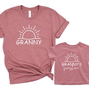 Może przedstawiać: Dwie koszulki w kolorze pudrowego r&oacute;żu. Większa koszulka z napisem "Granny" i grafiką słońca. Mniejsza koszulka z napisem "Granny's Sunshine" i grafiką słońca.