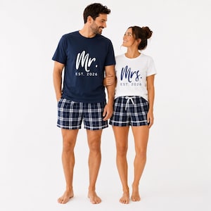 Pu&ograve; includere: Una t-shirt blu navy con scritta bianca Mr. EST. 2026 e una t-shirt bianca con scritta blu navy Mrs. EST. 2026. Entrambe le maglie sono abbinate a pantaloncini a quadri blu navy e bianchi con elastico in vita e cordoncino regolabile.