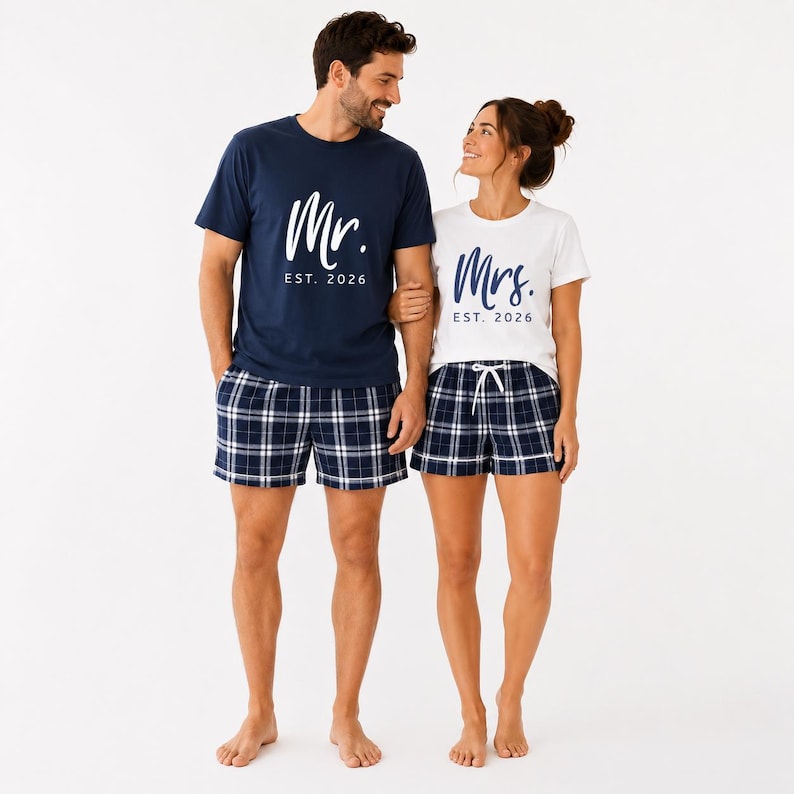 Pu&ograve; includere: Una t-shirt blu navy con scritta bianca Mr. EST. 2026 e una t-shirt bianca con scritta blu navy Mrs. EST. 2026. Entrambe le maglie sono abbinate a pantaloncini a quadri blu navy e bianchi con elastico in vita e cordoncino regolabile.