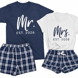 Pu&ograve; includere: Una t-shirt blu navy con testo bianco Mr. EST. 2026 e una t-shirt bianca con testo blu navy Mrs. EST. 2026, abbinate a due paia di pantaloncini a quadri blu navy, bianchi e neri con elastico in vita e cordoncino di chiusura.