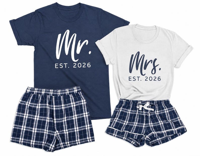 Pu&ograve; includere: Una t-shirt blu navy con testo bianco Mr. EST. 2026 e una t-shirt bianca con testo blu navy Mrs. EST. 2026, abbinate a due paia di pantaloncini a quadri blu navy, bianchi e neri con elastico in vita e cordoncino di chiusura.