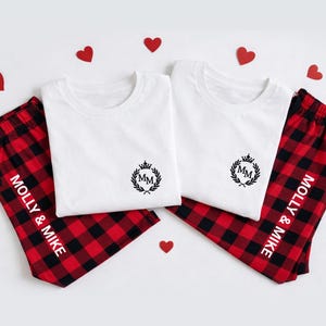 May include: Matching pajama set featuring white t-shirts with a black monogram and red and black plaid pajama pants. The pants are personalized with the names "Molly & Mike". Decorative red hearts are scattered around the set.
