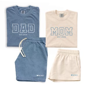 Peut inclure: Deux ensembles assortis de v&ecirc;tements d&eacute;contract&eacute;s. L'un est bleu avec "DAD EST. 2026" sur le t-shirt et le short avec "NOAH" et un c&oelig;ur. L'autre est cr&egrave;me avec "MOM EST. 2026" sur le t-shirt et le short avec "NOAH" et un c&oelig;ur.